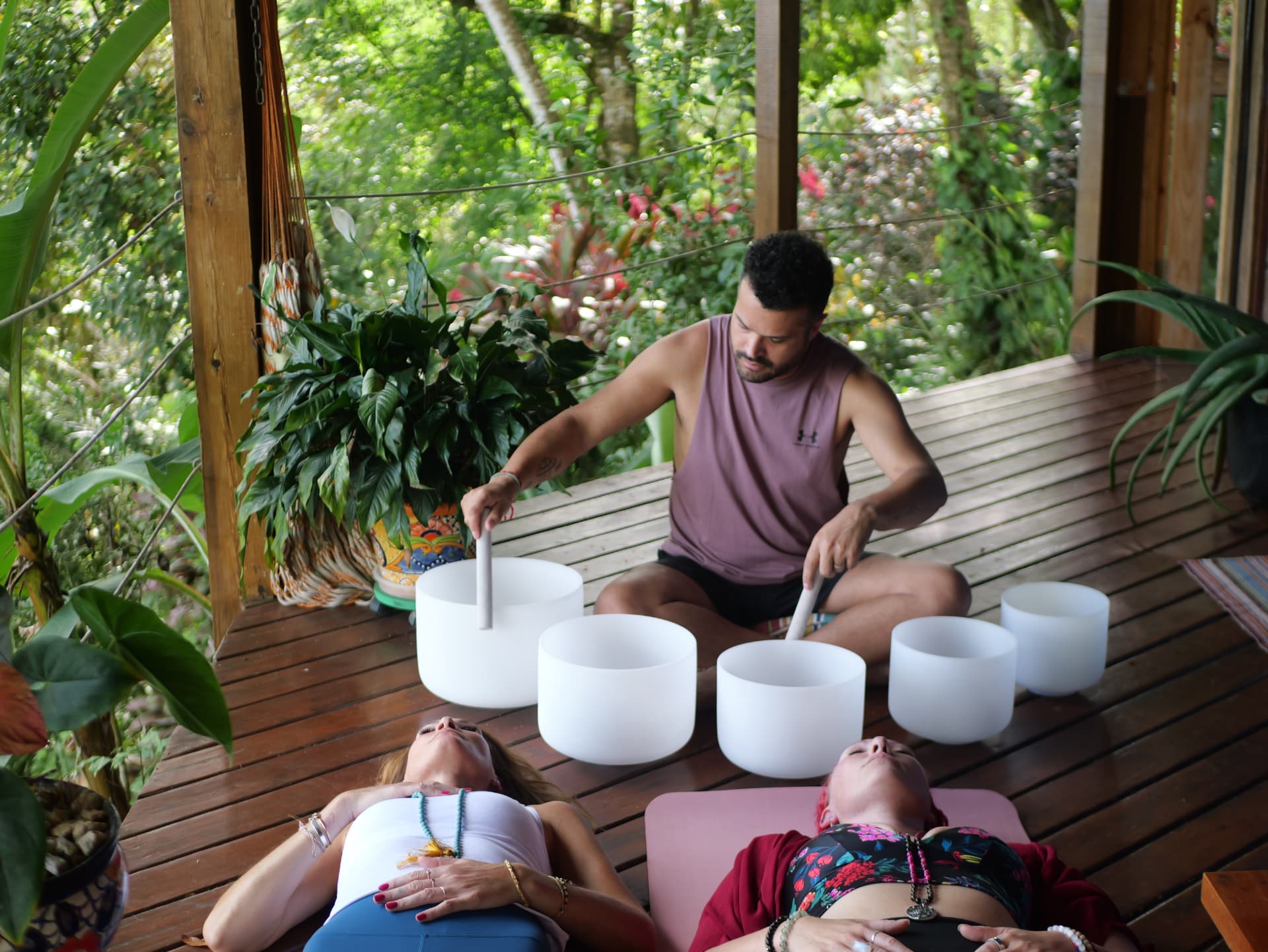 Sound healing practitioner playing crystal singing bowls for two people resting on a jungle deck